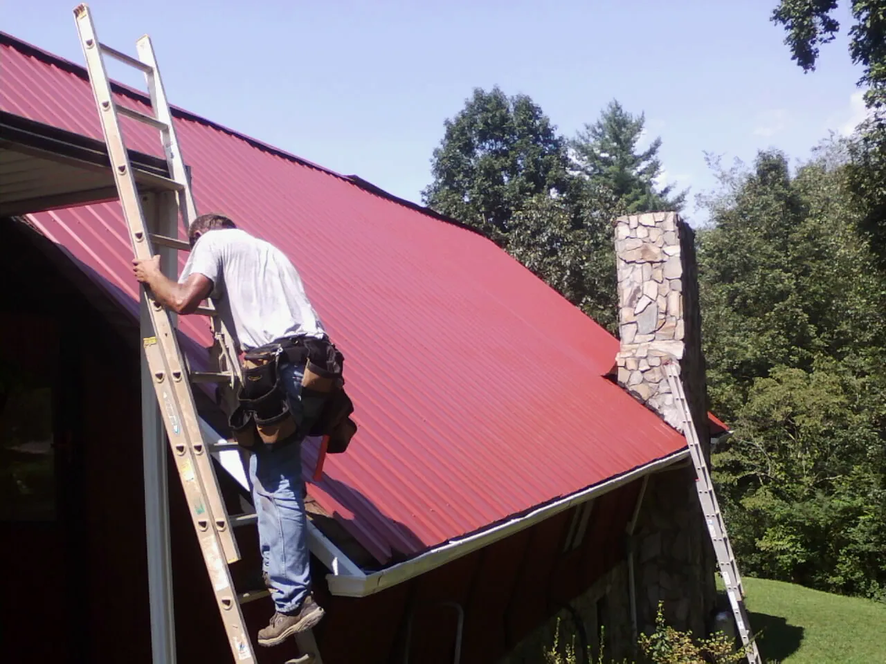 Roofer on a ladder beside a red metal roof and stone chimney — Soffit & Fascia Repair by Roofing Brothers in Chandlersville